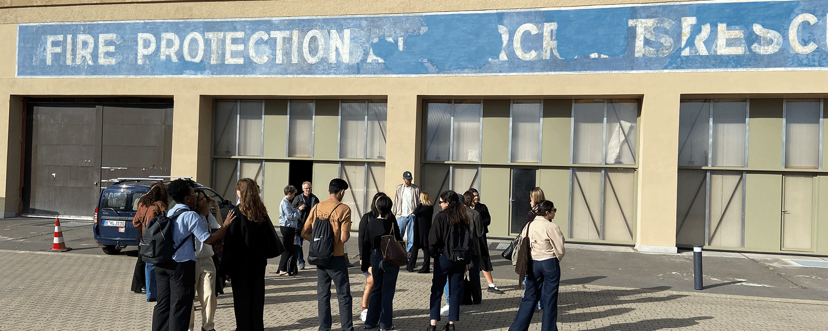 Eine Gruppe von Besuchenden steht vor der Alten Feuerwache auf dem Flughafen Tempelhof. Über mehreren Toren ist ein verblasstes blaues Wandbanner mit der Aufschrift „Fire Protection Workshop“ zu sehen. Die Personen versammeln sich auf einer gepflasterten Freifläche vor dem Gebäude.