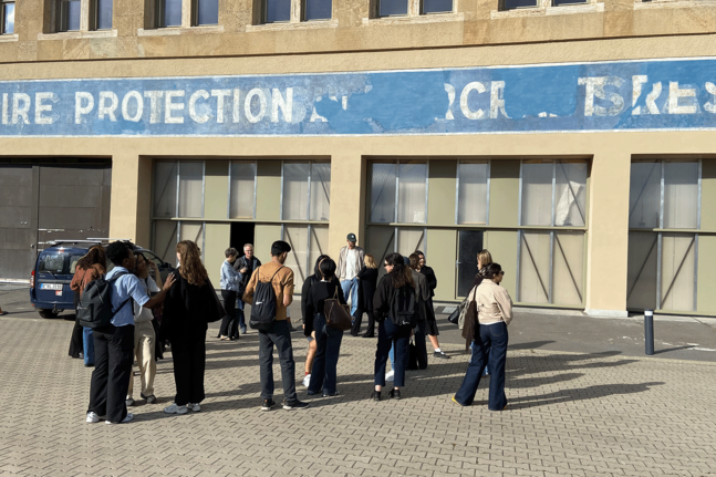 Eine Gruppe von Besuchenden steht vor der Alten Feuerwache auf dem Flughafen Tempelhof. Über mehreren Toren ist ein verblasstes blaues Wandbanner mit der Aufschrift „Fire Protection Workshop“ zu sehen. Die Personen versammeln sich auf einer gepflasterten Freifläche vor dem Gebäude.