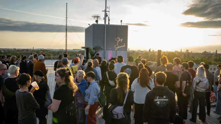 Eine große Menschenmenge steht auf einer Dachterrasse des THF TOWER bei Sonnenuntergang. Im Vordergrund sind zahlreiche Personen zu sehen, die in Richtung einer kleinen Bühne blicken, auf der eine Person spricht oder performt. Im Hintergrund erstreckt sich eine städtische Skyline unter einem warmen, leicht bewölkten Himmel.