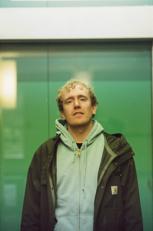 Carl Luis mit lockigem, blondem Haar steht vor einer grün getönten Glaswand; trägt einen hellblauen Hoodie unter einer dunklen Jacke mit sichtbarem Carhartt‑Logo.