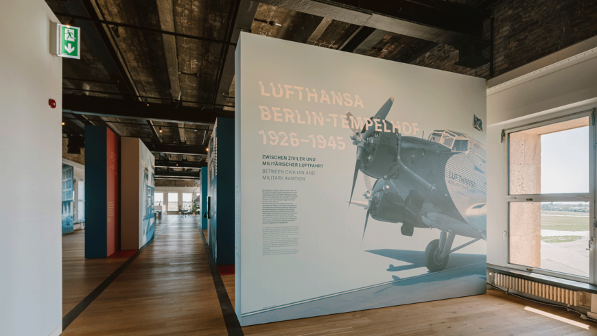 Lufthansa-Ausstellung mit großem Wandpanel zur Geschichte „Lufthansa Berlin‑Tempelhof 1926–1945“, darauf ein historisches Flugzeugfoto und zweisprachige Überschrift; im Hintergrund hohe Fenster mit Blick aufs Vorfeld sowie industriell anmutende Decke und Holzfußboden.