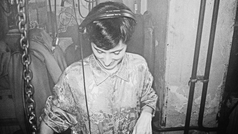  Schwarz-weiß-Porträt von DJ LEFEU, die mit Kopfhörern und konzentriertem Blick an einem DJ‑Equipment arbeitet. Sie trägt ein gemustertes Hemd mit hochgekrempelten Ärmeln. Im Hintergrund hängen Ketten, Kabel und industrielle Elemente, die eine rohe, atmosphärische Umgebung erzeugen.