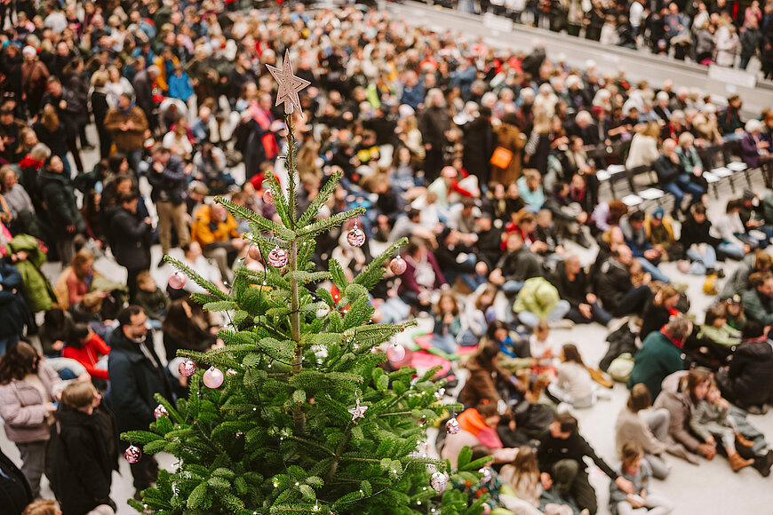 Menschenmenge sitzt auf dem Boden der Haupthalle am Flughafen Tempelhof. Im Vordergrund ein geschmückter Tannebaum
