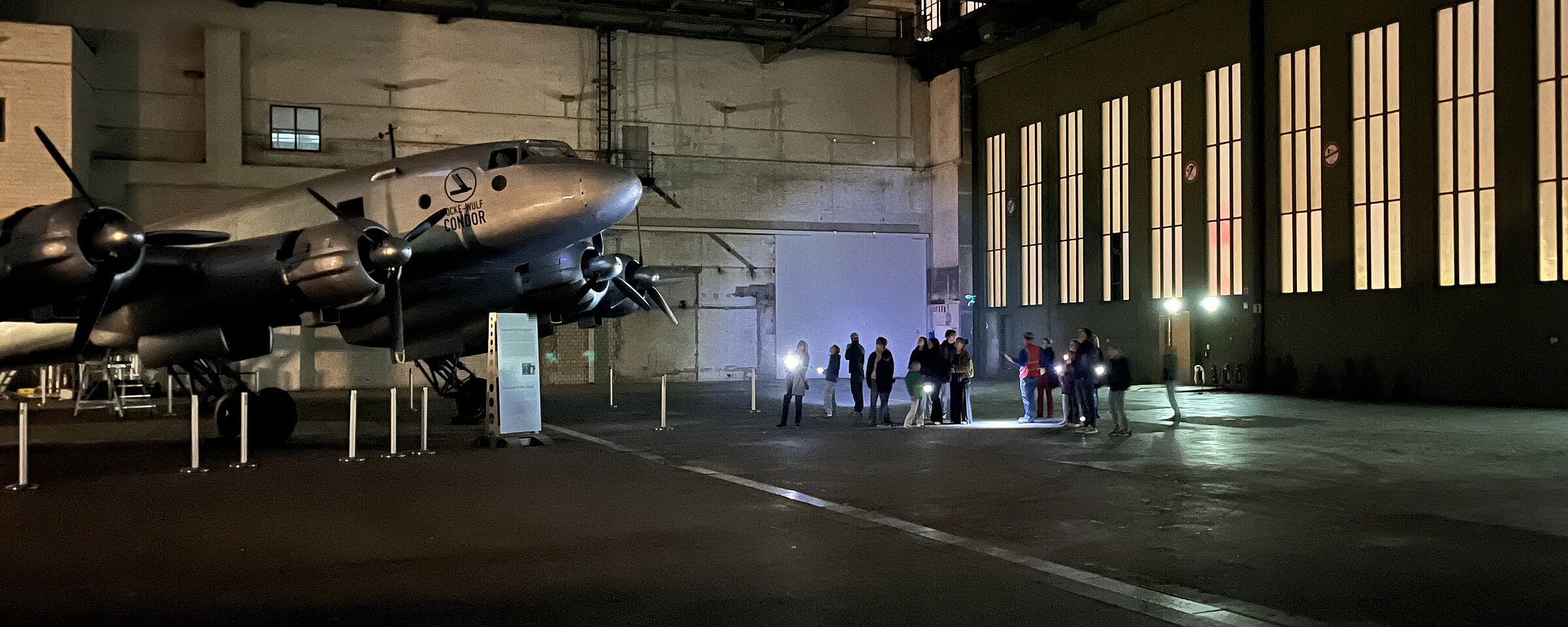 Kinder und Erwachsene leuchten bei einer Führung durch den Flughafen Tempelhof mit Taschenlampen auf ein Flugzeug, das in einem Hangar steht. 