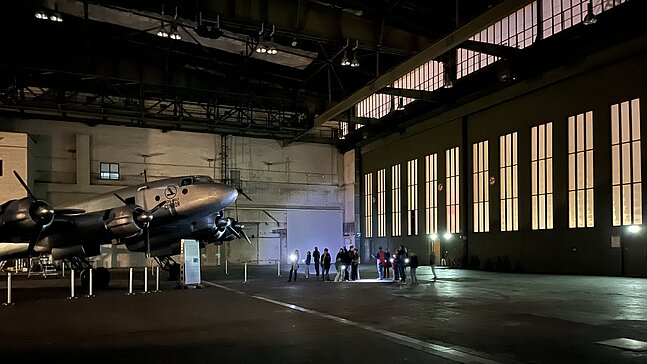 Kinder und Erwachsene leuchten bei einer Führung durch den Flughafen Tempelhof mit Taschenlampen auf ein Flugzeug, das in einem Hangar steht. 