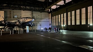 Kinder und Erwachsene leuchten bei einer Führung durch den Flughafen Tempelhof mit Taschenlampen auf ein Flugzeug, das in einem Hangar steht. 