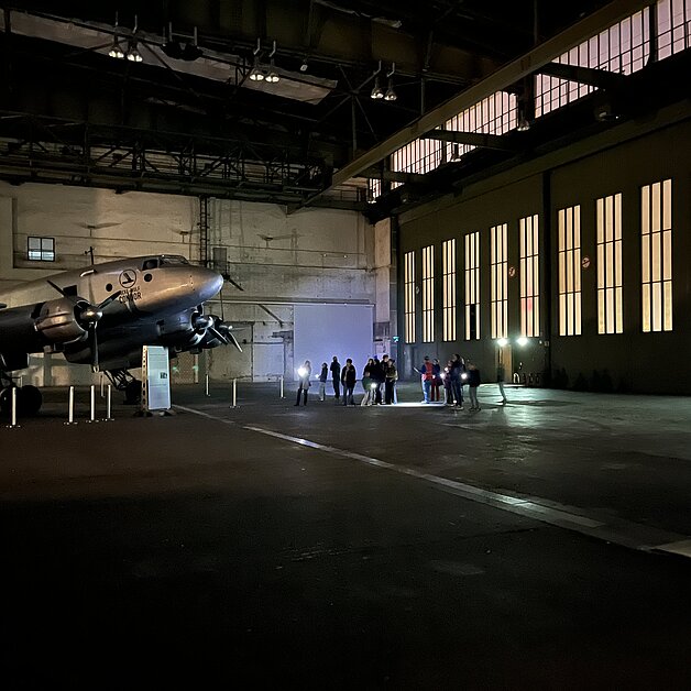 Kinder und Erwachsene leuchten bei einer Führung durch den Flughafen Tempelhof mit Taschenlampen auf ein Flugzeug, das in einem Hangar steht. 