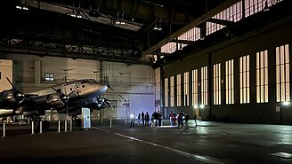 Kinder und Erwachsene leuchten bei einer Führung durch den Flughafen Tempelhof mit Taschenlampen auf ein Flugzeug, das in einem Hangar steht. 