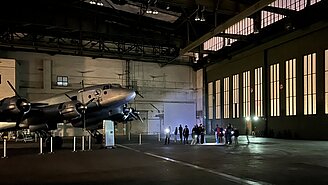 Kinder und Erwachsene leuchten bei einer Führung durch den Flughafen Tempelhof mit Taschenlampen auf ein Flugzeug, das in einem Hangar steht. 