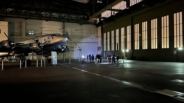 Kinder und Erwachsene leuchten bei einer Führung durch den Flughafen Tempelhof mit Taschenlampen auf ein Flugzeug, das in einem Hangar steht. 