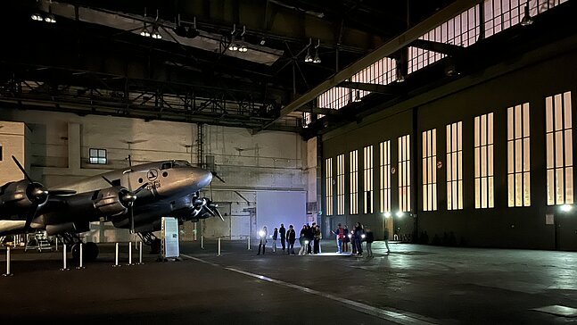 Kinder und Erwachsene leuchten bei einer Führung durch den Flughafen Tempelhof mit Taschenlampen auf ein Flugzeug, das in einem Hangar steht. 