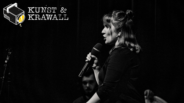 Ein dunkles Foto von einer Frau, die auf einer Bühne steht und in ihrer Hand ein Mikrophon hält. Oben links steht "Kunst &  Krawall" und ein gezeichneter Scheinwerfer
