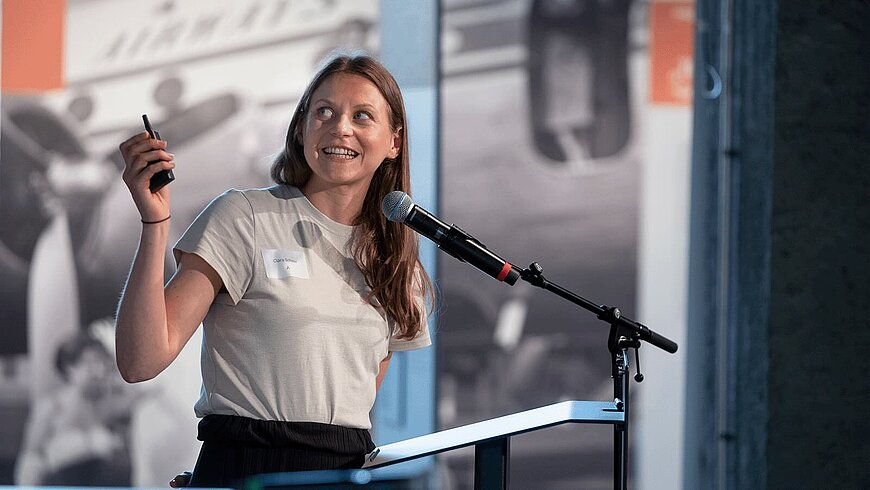 Eine Person steht an einem Rednerpult mit Mikrofon, lächelt und hält ein kleines Objekt in der Hand, während sie zu einem Publikum spricht. Im Hintergrund sind unscharfe Bilder oder Banner zu sehen.