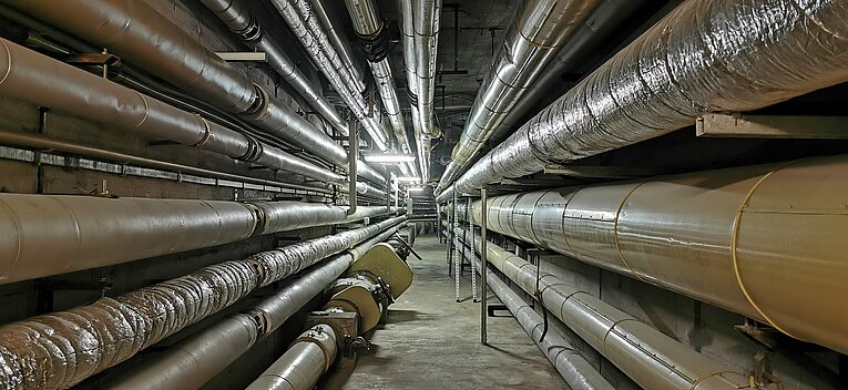 Ein langer, schmaler Versorgungsgang im Flughafen Tempelhof. An beiden Betonwänden und unter der Decke verlaufen zahlreiche dicke, isolierte Rohrleitungen parallel zueinander in die Ferne. Die Rohre sind teilweise mit silberner Folie oder beigen Schutzhüllen verkleidet.