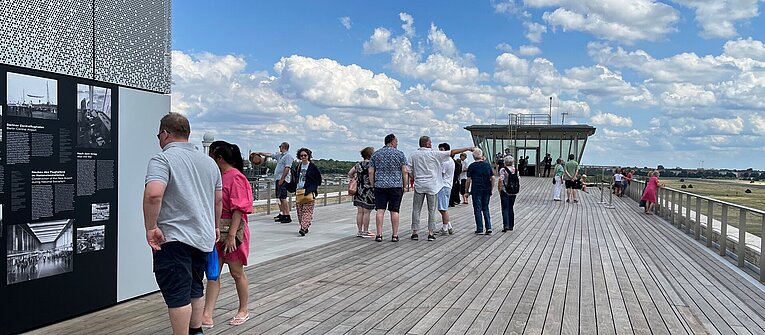 Menschen lesen sich auf der Dachterrasse des THF Towers Schilder durch. Im Hintergrund blauer Himmel