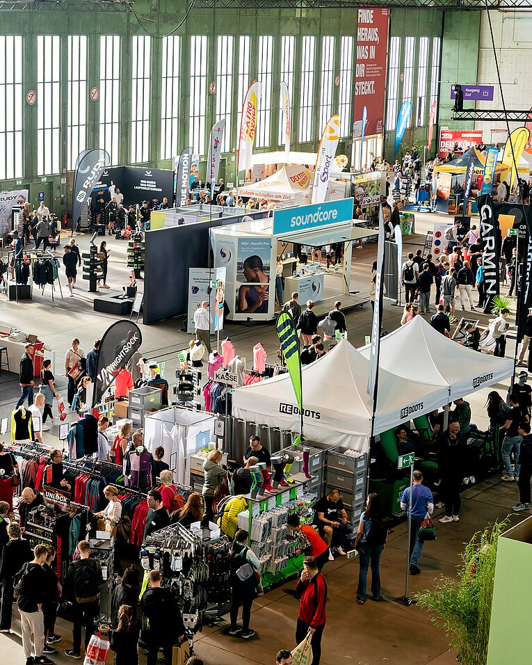 Vogelperspektive auf eine gut besuchte Messe im Hangar am Flughafen Tempelhof mit zahlreichen Informationsständen, Zelten und Bannern. Viele Menschen bewegen sich zwischen den Ständen in einem hellen, industriell gestalteten Gebäude mit großen Fensterfronten