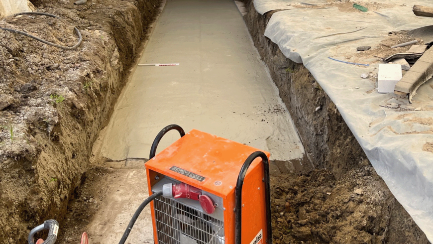 Schmale Baugrube mit frisch geglätteter Betonfläche, flankiert von abgedecktem Erdreich und Baumaterialien. Im Vordergrund steht ein orangefarmer Bautrockner mit Kabel, eingesetzt zur Trocknung oder Aushärtung des Betons.