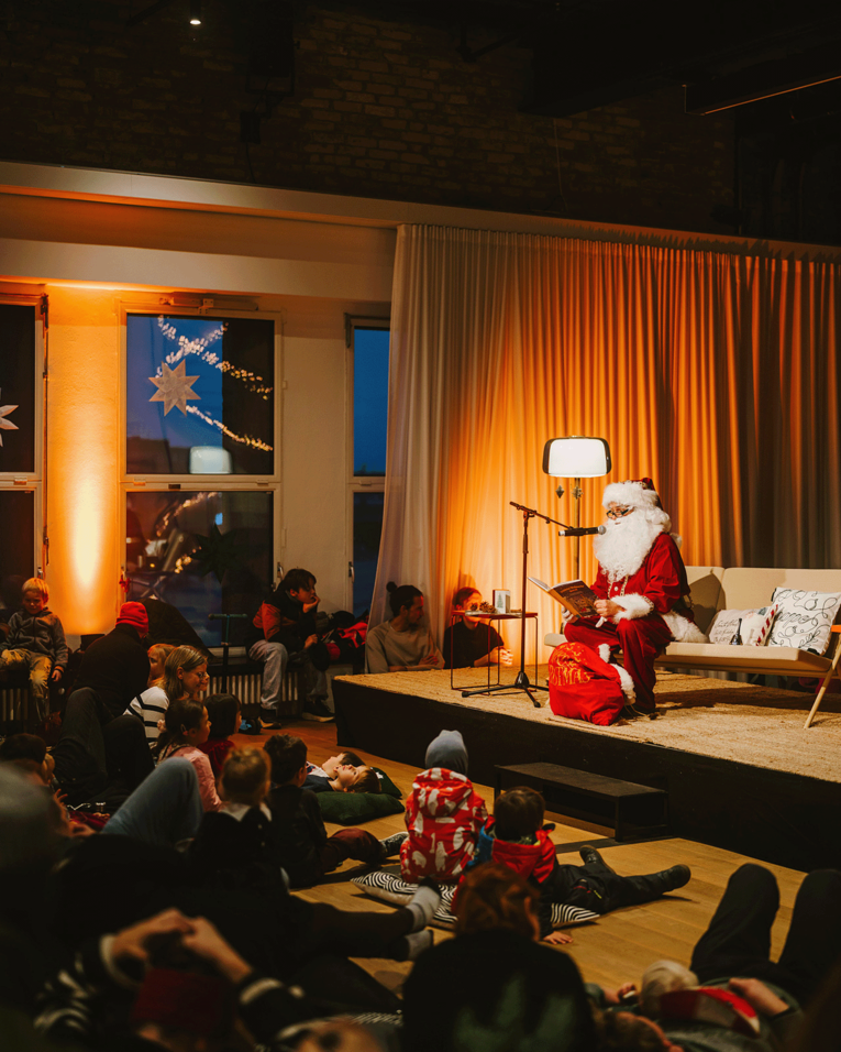 Beim Wintermarkt THF TOWER sitzt eine als Weihnachtsmann verkleidete Person auf einer kleinen Bühne und liest aus einem Buch vor. Zahlreiche Kinder und Erwachsene sitzen und liegen davor auf dem Boden und lauschen.