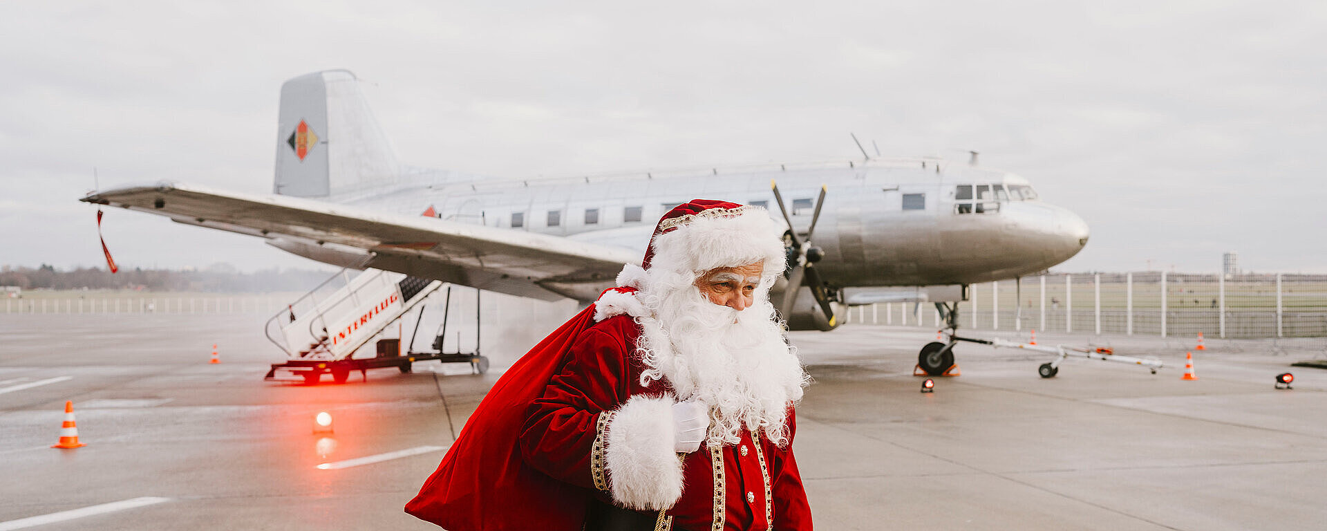 Ein verkleideter Weihnachtsmann mit einem großen roten Sack auf dem Rücken läuft am Flughafen Tempelhof vor einem Rosinenbomber-Flugzeug