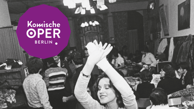 Historische Schwarz-Weiß-Aufnahme eines gut besuchten Gastraums mit mehreren an Tischen sitzenden und stehenden Personen. Im Vordergrund tanzt eine Person mit über dem Kopf erhobenen Armen zwischen den Tischen.