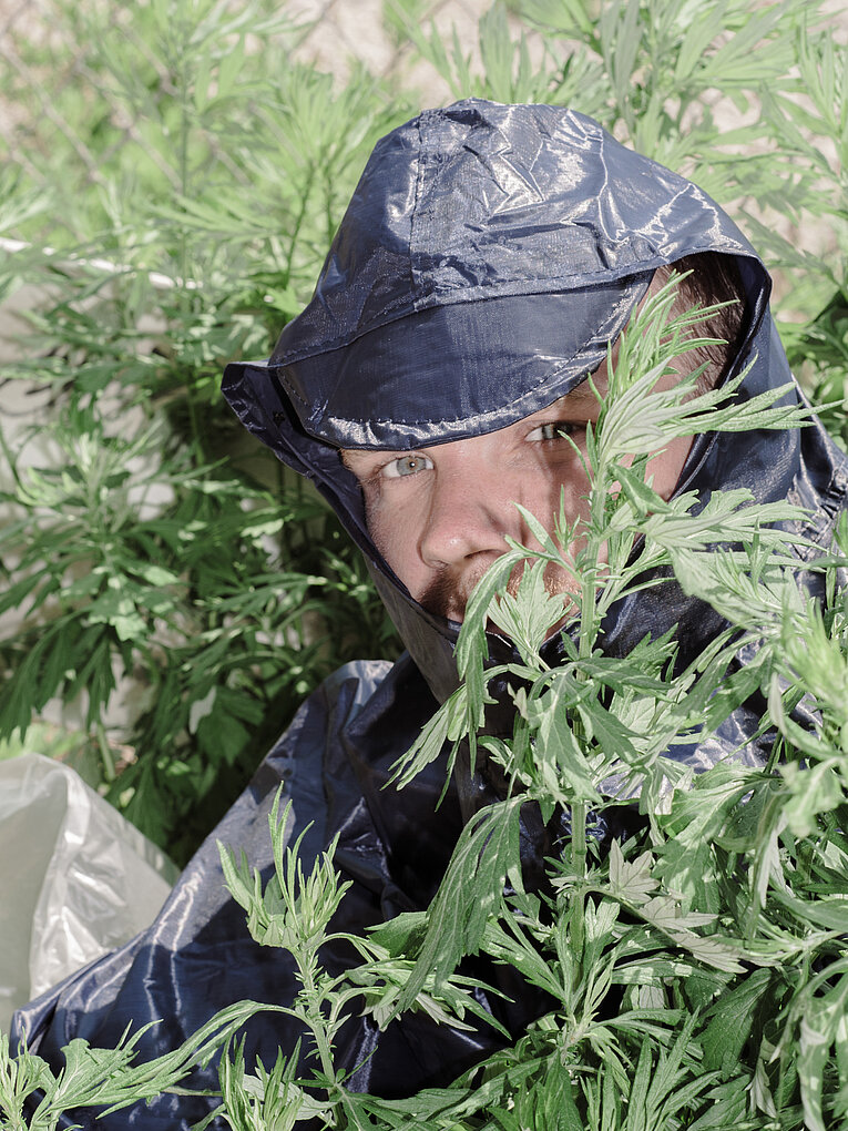 Justin Le Lover mit dunkelblauer Regenjacke steht zwischen dichtem grünen Blattwerk; das Gesicht ist teilweise von den Blättern umrahmt, wodurch ein Eindruck von Tarnung in der Vegetation entsteht.