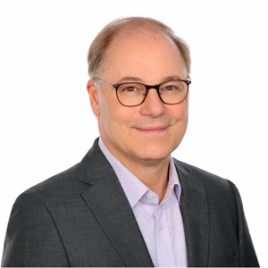 Porträt von Cornelius Grossmann mit Brille, hellem Hemd und dunklem Sakko vor weißem Hintergrund. Er lächelt leicht und blickt direkt in die Kamera. Das Foto ist professionell ausgeleuchtet und für Webnutzung optimiert.