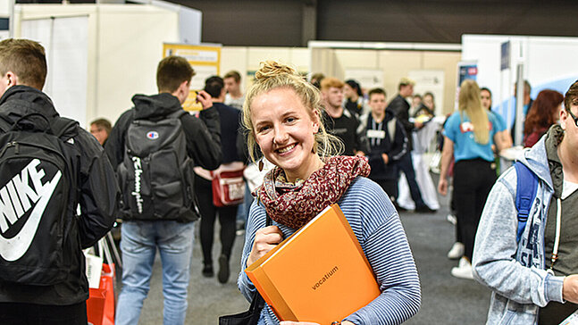 Junge Frau mit einem orangefarbenden Ordner auf der Messe vocatium blickt in die Kamera und lächelt.  Im Hintergrund sind mehrere junge Meschen zu sehen. 