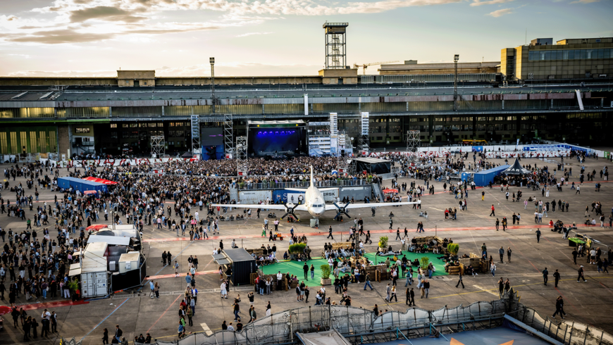Das TFELD Festival am Flughafen Tempelhof: Im Vordergrund steht ein zweimotoriges Propellerflugzeug, umgeben von Besucher*innen und mobilen Ständen. Dahinter versammelt sich eine dichte Menschenmenge vor einer Bühne mit blauem Licht und hohen Gerüsttürmen. Rundherum stehen Container, Zelte und ein „BAR“-Stand. Im Hintergrund erhebt sich das Flughafen Gebäude mit Turm, unter einem Himmel mit warmem Sonnenlicht.
