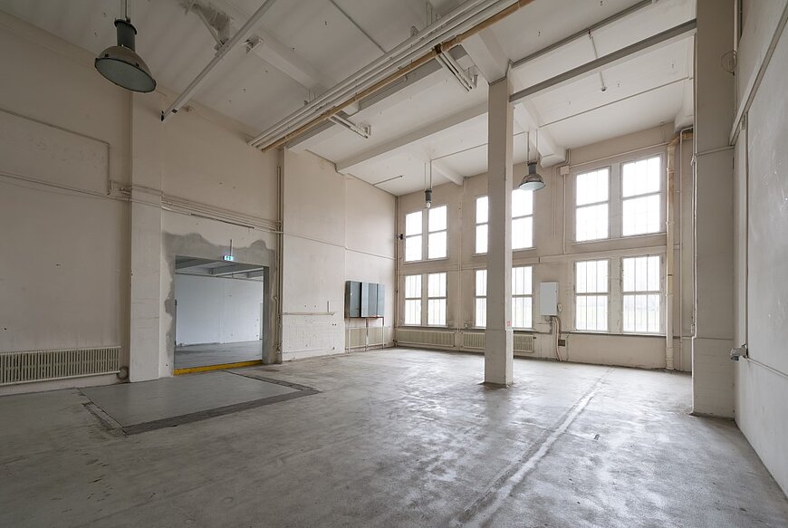 Großer, leerer, heller Showroom Hangar 6 mit hellgrauem Betonboden. Hohe Wände, weiße Deckenbalken, zwei massive Säulen und große Fenster mit Sprossen, die viel Tageslicht hereinlassen. Links ist ein breiter Durchgang zu einem weiteren Raum.