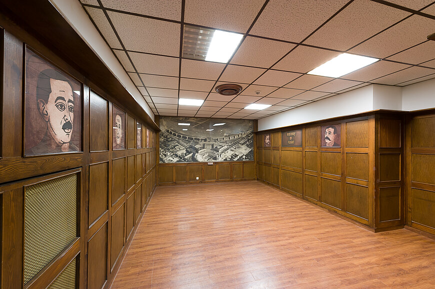 Langer, leerer Raum mit holzvertäfelten Wänden und heller Rasterdecke. An den Wänden hängen Porträts gezeichneter Gesichter. Am Ende des Raumes ist ein großes, sepiafarbenes Wandbild einer historischen Luftaufnahme vom Flughafen Tempelhof angebracht. Der Boden besteht aus hellem Holz