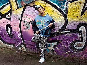 Ein Mann steht vor eine mit Graffiti besprühten Wand und hält eine Querflöte in der Hand.