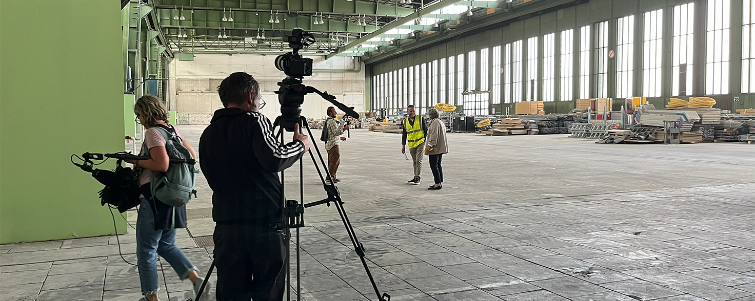 Dreharbeiten in einer riesigen, leeren einem Flugzeughangar am Flughafen Tempelhof. Im Vordergrund sind drei Personen aus dem Aufnahmeteam zu sehen, darunter eine Person von hinten, die eine professionelle Kamera auf einem Stativ bedient. Zwei weitere Personen (eine mit einer Videokamera über der Schulter und eine, die ihnen gegenübersteht) blicken in die Tiefe der Halle. Der massive Raum zeichnet sich durch eine hohe Decke mit Stahlträgern, große Fensterfronten auf der rechten Seite und einen grauen Betonboden aus. Im Hintergrund sind aufgestapelte Baumaterialien und Paletten zu erkennen.