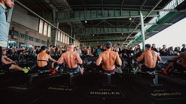 Sechs shirtlose Athleten sitzen nebeneinander auf Ruderergometern und treten in einem Wettkampf an. Hinter ihnen steht eine Menschenmenge, die durch Absperrungen getrennt zuschaut. Die Halle wirkt industriell mit hohen Decken und sichtbaren Trägern.