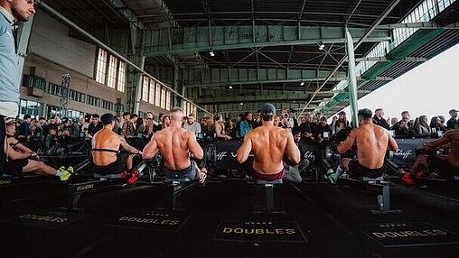 Sechs shirtlose Athleten sitzen nebeneinander auf Ruderergometern und treten in einem Wettkampf an. Hinter ihnen steht eine Menschenmenge, die durch Absperrungen getrennt zuschaut. Die Halle wirkt industriell mit hohen Decken und sichtbaren Trägern.