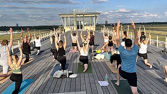 Auf dem Foto zu sehen ist die lange hölzerne Dachterrasse und Menschen, die mit dem Rücken zur Kamera in einer Yogapose ausharren.