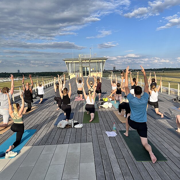 Auf dem Foto zu sehen ist die lange hölzerne Dachterrasse und Menschen, die mit dem Rücken zur Kamera in einer Yogapose ausharren.