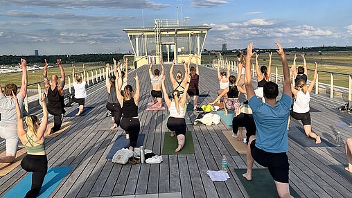 Auf dem Foto zu sehen ist die lange hölzerne Dachterrasse und Menschen, die mit dem Rücken zur Kamera in einer Yogapose ausharren.