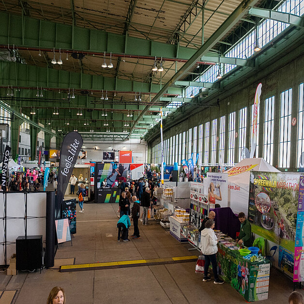 Blick in den Hangar am Flughafen Tempelhof, in dem während der Messe zum Halbmarathon Stände und Flaggen aufgestellt sind. Menschen stehen herum und schauen sich die Stände an
