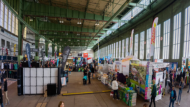 Blick in den Hangar am Flughafen Tempelhof, in dem während der Messe zum Halbmarathon Stände und Flaggen aufgestellt sind. Menschen stehen herum und schauen sich die Stände an
