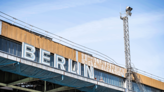 Schriftzug BERLIN an der Dachfassade des Flughafen Tempelhof.  Rechts daneben steht die Höhenangabe „164 FEET FIELD ELEVATION“. Unter der Dachkante ist ein Metallgerüst mit Netzstruktur sichtbar, darüber ein hoher Mast vor blauem Himmel.