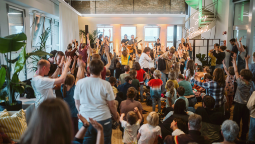 Kinder und Familien bei der Ferienstartparty  im THF TOWER mit hohen Decken und großen Fenstern; viele sitzen, andere stehen und heben die Hand, als Teil eines interaktiven Workshops oder Meetings.