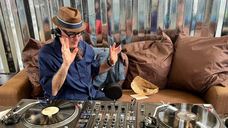 DJ Olli Goolightly mit braunem Hut, blauer Binde und Kopfhörern sitzt auf einem braunen Sofa vor einer metallischen Wand und bedient DJ‑Equipment mit zwei Plattenspielern und Mischpult; entspannte Indoor‑Szene mit professionellem Musik‑Setup.