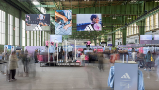 Große Marathon‑Expo in einem Hangar am Flughafen Tempelhof mit hohen Decken und grünen Stahlträgern; Adidas‑Branding und Banner mit Läufermotiven hängen von oben. Im Zentrum stehen Sport‑Mannequins in Laufbekleidung, umgeben von farblich sortierten Kleiderständern.