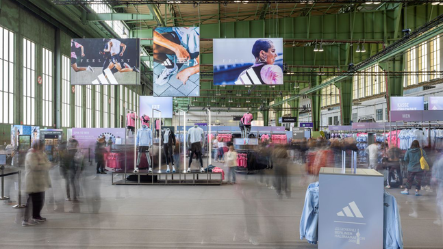 Große Marathon‑Expo in einem Hangar am Flughafen Tempelhof mit hohen Decken und grünen Stahlträgern; Adidas‑Branding und Banner mit Läufermotiven hängen von oben. Im Zentrum stehen Sport‑Mannequins in Laufbekleidung, umgeben von farblich sortierten Kleiderständern.
