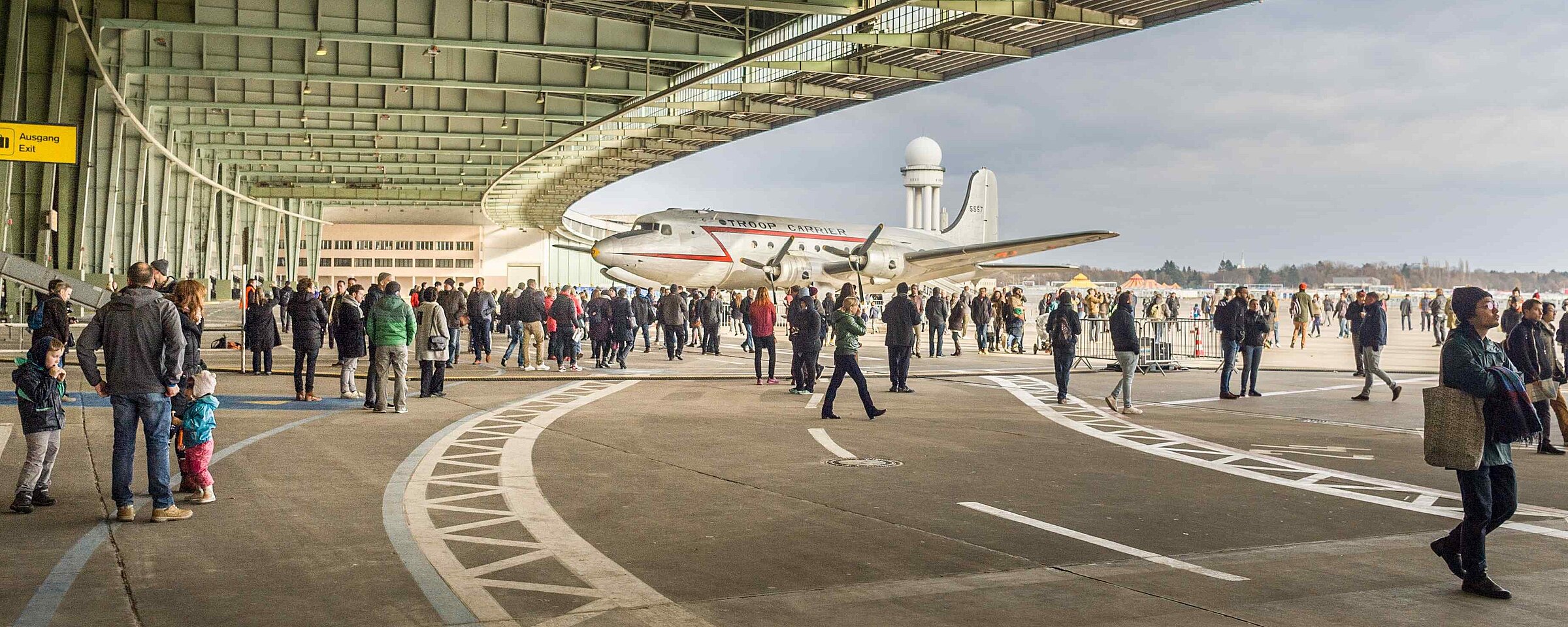 Menschenmenge bei einem Event am Flughafen Tempelhof unter einem modernen, überdachten Bereich mit offener Seitenansicht und Blick auf den Himmel.