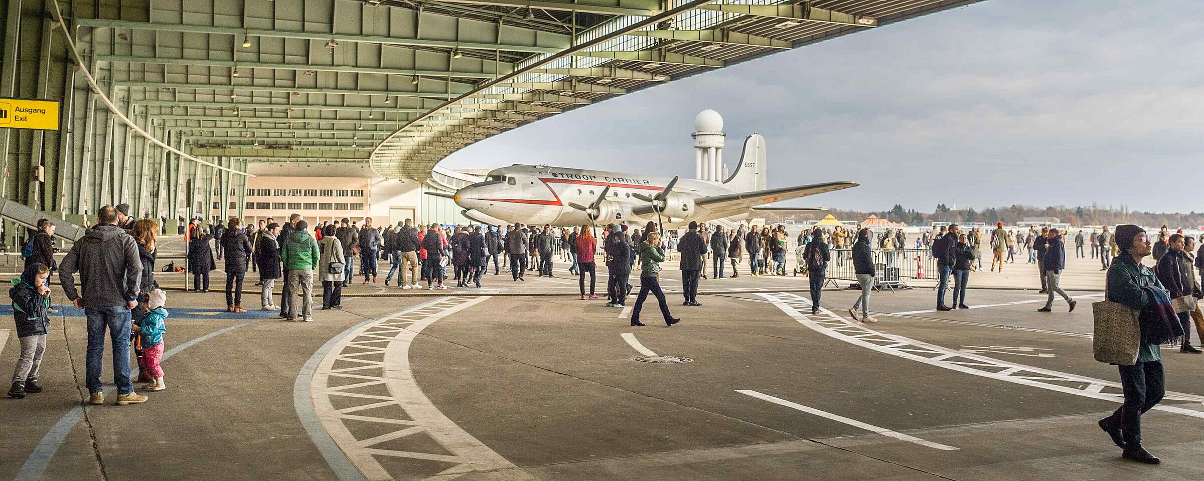 Menschenmenge bei einem Event am Flughafen Tempelhof unter einem modernen, überdachten Bereich mit offener Seitenansicht und Blick auf den Himmel.