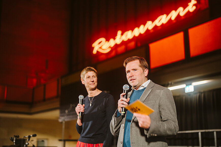 Geschäftsführer Fabian Schmitz-Grethlein und Pressesprecherin Nina Mützen stehen auf einer Bühne. Bei halten ein Mikrophon in der Hand. Im Hintergrund der rot leuchtende Schriftzug "Restaurant"