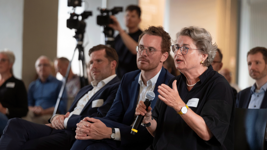 Foto von Petra Kahlfeldt, die in ein Mikrophon spricht. neben ihr sitzt Christoph Rauhut und hinter ihm Fabian Schmitz-Grethlein. Im Hintergrund steht ein Kameramann und Publikum ist zu sehen