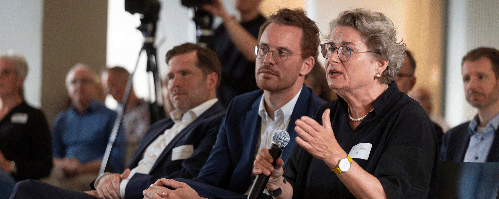 Foto von Petra Kahlfeldt, die in ein Mikrophon spricht. neben ihr sitzt Christoph Rauhut und hinter ihm Fabian Schmitz-Grethlein. Im Hintergrund steht ein Kameramann und Publikum ist zu sehen