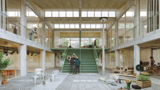 Entwurf eines Innenraums mit breiten grünen Stufen, hohen Glasfronten und sichtbaren Betonstrukturen. Menschen bewegen sich auf beiden Ebenen, während im Erdgeschoss verstreute Stühle, Pflanzen, Werkzeuge und Materialien eine Mischung aus Arbeits‑ und Gemeinschaftsbereich erkennen lassen.